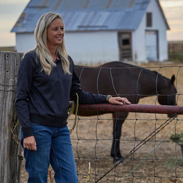 A woman, Shali Lord, standing and posing at a fence next to her horse looking away from the camera.